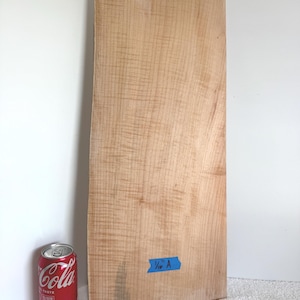 May include: A large, light-coloured piece of wood with a wavy grain pattern. A red Coca-Cola can is next to the wood for scale. A blue piece of tape with the text "1/4 A" is affixed to the wood. The wood appears to be a slab or plank.