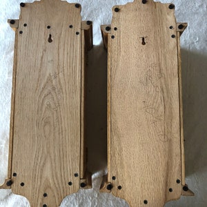 K&ouml;nnte beinhalten: Zwei Holz-Wandregale mit einem dekorativen oberen Rand und schwarzen Metallschrauben. Die Regale sind aus hellem braunem Holz gefertigt und haben ein Schl&uuml;sselloch auf der R&uuml;ckseite.