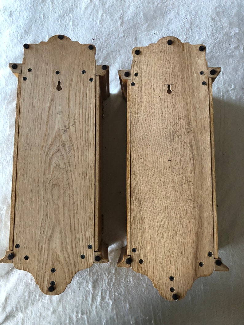 K&ouml;nnte beinhalten: Zwei Holz-Wandregale mit einem dekorativen oberen Rand und schwarzen Metallschrauben. Die Regale sind aus hellem braunem Holz gefertigt und haben ein Schl&uuml;sselloch auf der R&uuml;ckseite.