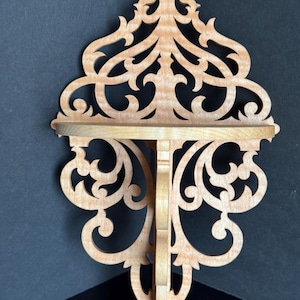 Op de afbeelding: Een houten hoekplank met ingewikkelde, lasergesneden decoratieve details. De plank is gemaakt van lichtbruin hout en heeft een gegolfde rand.