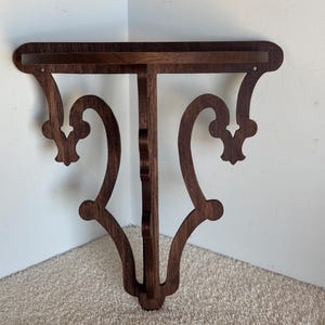 Puede incluir: Un estante de esquina de madera marrón oscuro con intrincados detalles de volutas. El estante está diseñado para ser montado en una pared.