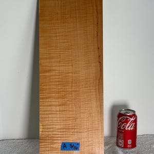 May include: A rectangular piece of light brown wood with a wavy grain pattern. A blue piece of tape with the text "A 5/16" is affixed to the bottom. A red Coca-Cola can is next to the wood.