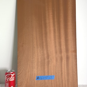 Puede incluir: Una pieza rectangular de chapa de madera de color marrón rojizo con un patrón de veta visible. Una cinta azul está pegada a la superficie con el texto A-1/16" x 14 3/4" x 24". Una lata de refresco roja está junto a la madera como referencia.
