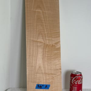 May include: A rectangular piece of light-colored wood with visible grain patterns. A blue tape with the text "3/8" A" is affixed to the wood. A red Coca-Cola can is placed next to the wood for size comparison.