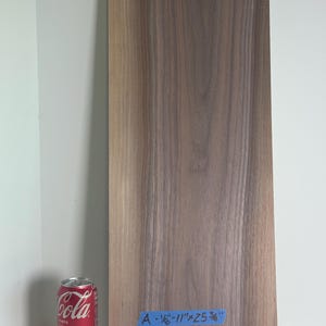 May include: A rectangular brown wooden board with a visible wood grain pattern. A blue piece of tape is attached to the bottom with the text A - 1 1/8" x 11" x 25 3/8" written in black ink. A red soda can is positioned to the left of the board.