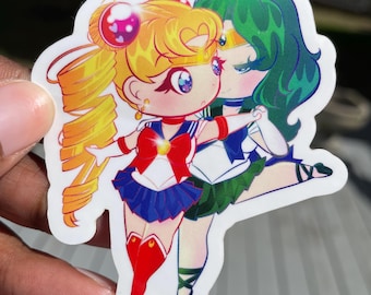 Puede incluir: Una pegatina con una ilustración de estilo chibi de dos personajes de anime. El personaje de la izquierda lleva un uniforme de marinero azul y rojo con un lazo amarillo y una estrella en el pecho. El personaje de la derecha lleva un uniforme de marinero verde y blanco con un lazo verde y una estrella en el pecho. Ambos personajes tienen ojos grandes y sonríen.