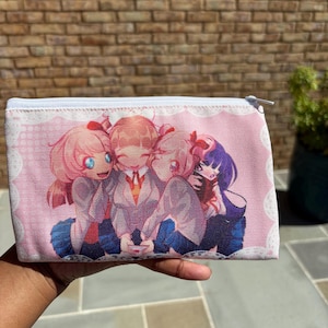 Peut inclure: Une pochette rectangulaire rose avec une fermeture éclair et une illustration de style anime de quatre personnages en uniformes scolaires. La pochette a une fermeture éclair blanche et une bordure en dentelle blanche.