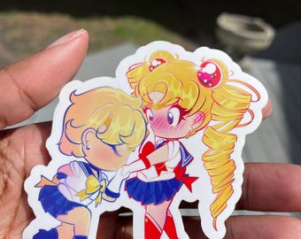 Puede incluir: Una pegatina con dos personajes de anime, uno con cabello rubio y un atuendo azul y amarillo, el otro con cabello rubio y un atuendo azul y rojo. Los personajes están en una pose romántica, con el personaje con el atuendo azul y amarillo sosteniendo la mano del otro personaje.