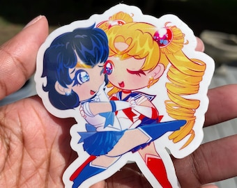 Puede incluir: Una pegatina con una ilustración de dibujos animados de dos personajes de anime, uno con cabello azul y el otro con cabello amarillo, ambos con atuendos de marinero. Los personajes se abrazan y sonríen.