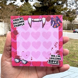 Könnte beinhalten: Ein quadratischer rosa Notizblock mit einem Herzmuster und dem Text "YOU'RE KILLING PEOPLE" und "NO, I'M KILLING BOYS". Der Notizblock hat Vampirzähne, eine lila Jacke und Brillenaufkleber.