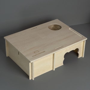 Op de afbeelding: Een lichtgekleurd houten hamsterhuis met een cirkelvormige opening en een decoratieve boogvormige ingang. De tekst "HAMSTERHOME" en een berenlogo zijn zichtbaar op het bovenoppervlak. De structuur is rechthoekig en lijkt van multiplex te zijn.