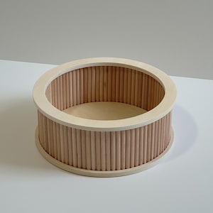 Può includere: Un contenitore rotondo in legno chiaro con un bordo e tasselli di legno verticali che formano i lati. La base interna è di una tonalità di legno più chiara. Il contenitore è probabilmente per lo stoccaggio o l'esposizione.