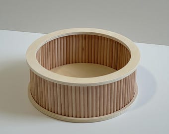 May include: A round, light-colored wooden container with a rim and vertical wooden dowels forming the sides. The interior base is a lighter shade of wood. The container is likely for storage or display.