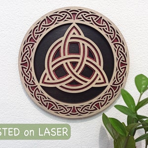 May include: A circular wooden wall decor with a red and black Celtic knot design. The knot is a triquetra, a symbol of the trinity. The design is intricate and detailed, and the wood is polished to a smooth finish. "TESTED on LASER" is printed in green at the bottom of the image.