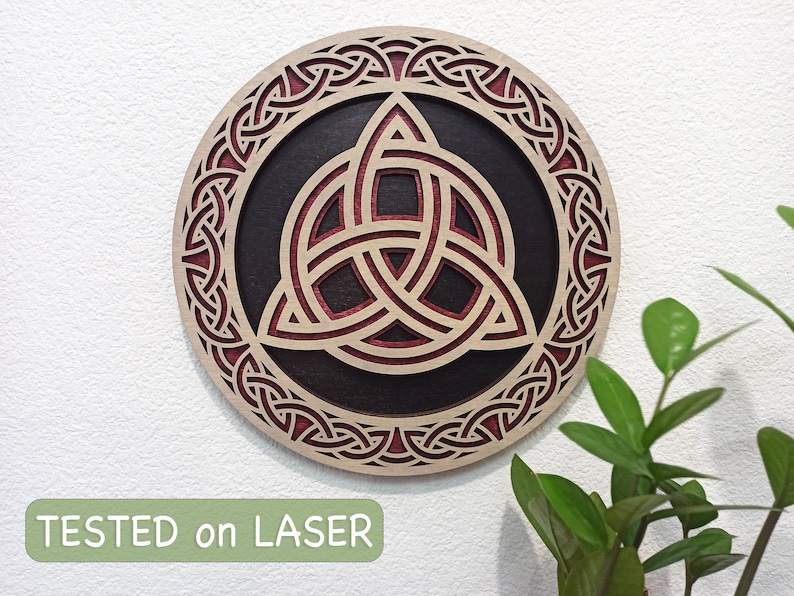 May include: A circular wooden wall decor with a red and black Celtic knot design. The knot is a triquetra, a symbol of the trinity. The design is intricate and detailed, and the wood is polished to a smooth finish. "TESTED on LASER" is printed in green at the bottom of the image.