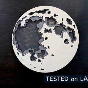 May include: A white wooden moon with a black and gray detailed design. The moon is cut out in a layered style, creating a 3D effect. The text "TESTED on LASER" is printed below the moon.