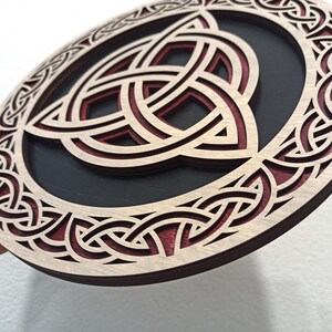 May include: A circular wooden wall decor with a red and brown Celtic knot design. The knot is a triple knot, also known as a trinity knot, and is a symbol of unity, strength, and eternity.