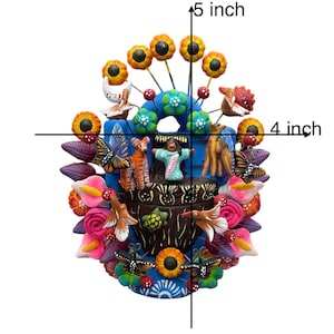 May include: A colorful ceramic wall hanging featuring a blue pot with a brown rim, surrounded by flowers, butterflies, birds, and animals. The pot is decorated with a green turtle and a small figure in a blue dress. The wall hanging measures 4 inches wide and 5 inches tall.