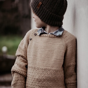Puede incluir: Una persona joven con un gorro de punto marrón y un suéter de punto beige con un detalle de botón en el hombro.