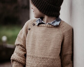 May include: A young person wearing a brown knitted beanie and a beige knitted sweater with a button detail on the shoulder.