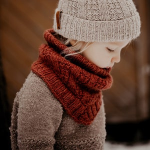 May include: A young child wearing a beige knitted beanie with a brown leather strap and a rust-coloured knitted scarf. The child is looking down and the background is blurred.
