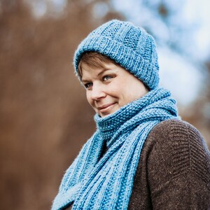 May include: A woman wearing a blue knitted hat and matching scarf. The hat and scarf are both made with a textured stitch.