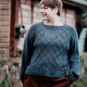 Könnte beinhalten: Ein blauer Strickpullover mit Rautenmuster. Der Pullover hat lange Ärmel und einen Rundhalsausschnitt. Die Person trägt eine braune Hose. Der Hintergrund umfasst ein Backsteingebäude und Grün.