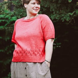 Könnte beinhalten: Ein korallenfarbenes Strickoberteil mit kurzen Ärmeln und einem dekorativen Saum. Das Oberteil wird mit einem schwarz-weiß karierten Rock kombiniert. Das Outfit wird von einer Person vor einem grünen, belaubten Hintergrund getragen.
