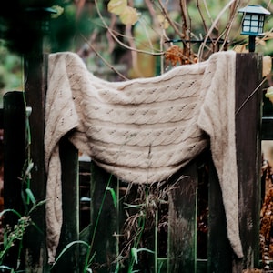Könnte beinhalten: Ein beigefarbener Strickschal, der über einem verwitterten Holzzaun drapiert ist. Der Schal hat ein Zopfmuster und besteht aus einem weichen, warmen Material. Der Zaun ist dunkelbraun und von Grün umgeben.