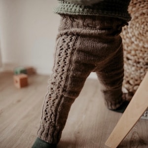 Może przedstawiać: Brązowe dzianinowe legginsy dla niemowląt z wzorem warkocza. Legginsy nosi niemowlę stojące w pobliżu drewnianej zabawki.