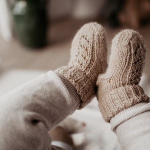 Pode incluir: Um par de botinhas de bebé de malha bege com um delicado padrão de renda. As botinhas são usadas por um bebé com os pés cruzados.