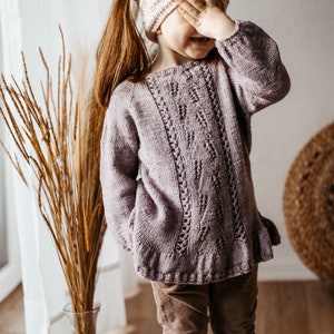 Può includere: Una bambina che indossa un maglione di lana viola con un motivo a treccia e pantaloni di velluto a coste marroni. Indossa una fascia per capelli color crema e ha la mano sugli occhi.