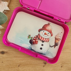 Könnte beinhalten: Rosa Lunchbox mit einem Schneemann, der ein Eis am Stiel hält. Der Schneemann trägt einen roten Hut und einen rot-weiß gestreiften Schal. Die Lunchbox hat ein herausnehmbares Tablett mit Fächern.