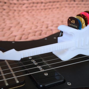 以下が含まれることがあります： ジム・ダンロップのロゴが付いた白いプラスチック製のギター型のピックホルダーで、さまざまな色のギターピックが入っています。ホルダーは黒いエレキギターの上に置かれています。