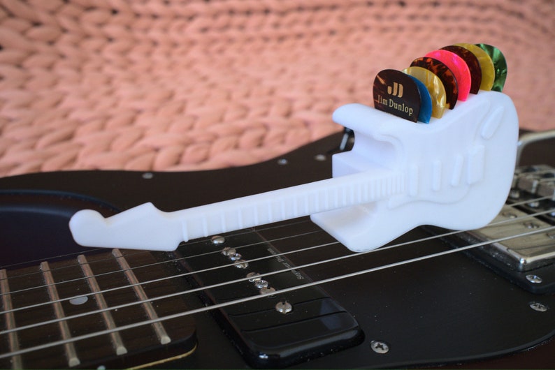 以下が含まれることがあります： ジム・ダンロップのロゴが付いた白いプラスチック製のギター型のピックホルダーで、さまざまな色のギターピックが入っています。ホルダーは黒いエレキギターの上に置かれています。