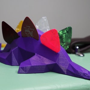 May include: Purple 3D printed guitar pick holder shaped like a dinosaur with five guitar picks in various colors and materials, including red, green, yellow, and clear.