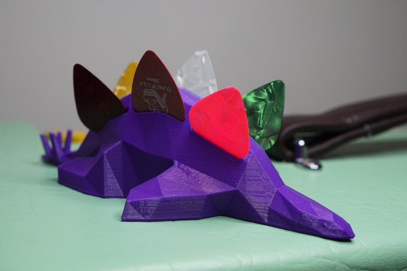May include: Purple 3D printed guitar pick holder shaped like a dinosaur with five guitar picks in various colors and materials, including red, green, yellow, and clear.