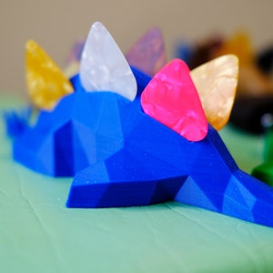 May include: Blue 3D printed dinosaur figurine with colorful guitar picks on its back. The dinosaur is on a green surface.