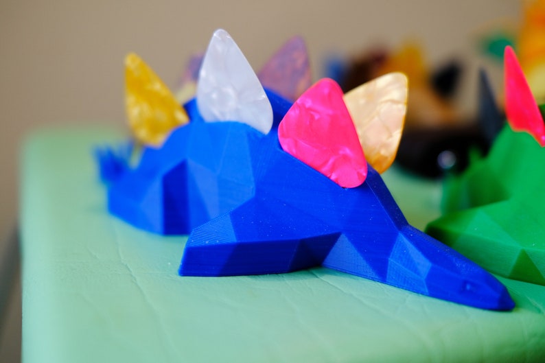 May include: Blue 3D printed dinosaur figurine with colorful guitar picks on its back. The dinosaur is on a green surface.