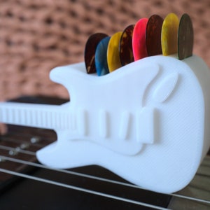 以下が含まれることがあります： 白いプラスチック製のギター型のピックホルダーに、カラフルなギターピックが並んでいます。ホルダーは黒いギターの上に置かれています。