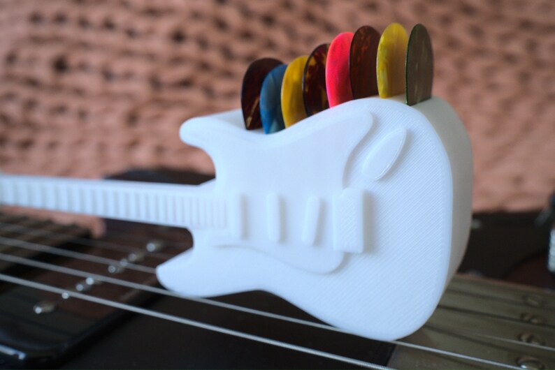 以下が含まれることがあります： 白いプラスチック製のギター型のピックホルダーに、カラフルなギターピックが並んでいます。ホルダーは黒いギターの上に置かれています。