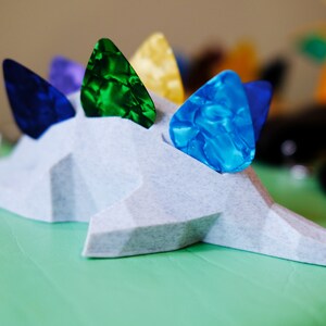May include: A white, 3D printed dinosaur-shaped guitar pick holder with five guitar picks in various colors: blue, green, yellow, purple, and blue.