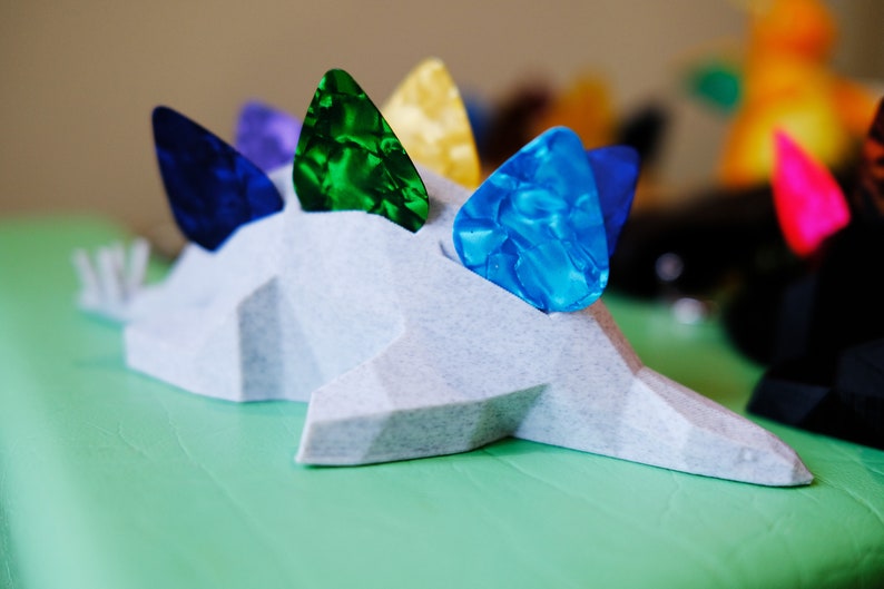 May include: A white, 3D printed dinosaur-shaped guitar pick holder with five guitar picks in various colors: blue, green, yellow, purple, and blue.