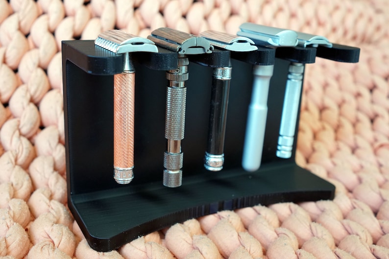 May include: A black plastic stand with five silver safety razors. The razors have different colored handles: copper, silver, black, and gray.
