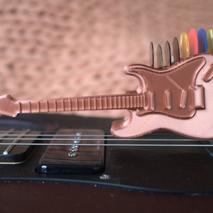 以下が含まれることがあります： 背面にピックホルダーが付いた、小さな銅色の3Dプリントギター。ギターは黒いエレキギターの上に置かれています。