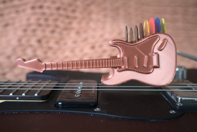以下が含まれることがあります： 背面にピックホルダーが付いた、小さな銅色の3Dプリントギター。ギターは黒いエレキギターの上に置かれています。