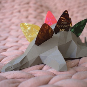 May include: A gray, geometric dinosaur-shaped guitar pick holder with five guitar picks inserted. The picks are yellow, brown, pink, and green.