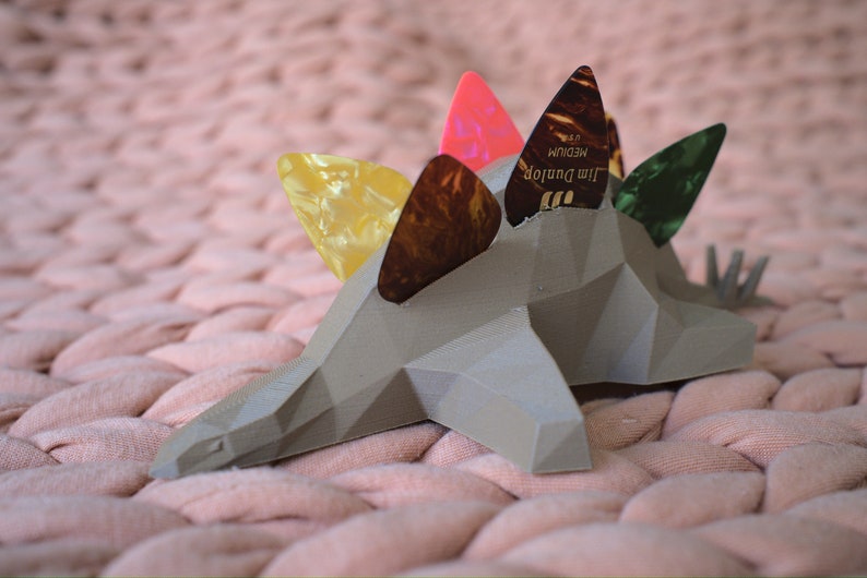 May include: A gray, geometric dinosaur-shaped guitar pick holder with five guitar picks inserted. The picks are yellow, brown, pink, and green.
