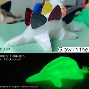 May include: A white 3D printed dinosaur figurine with a glow-in-the-dark finish. The dinosaur is holding guitar picks in its back spikes. The dinosaur glows green in the dark after being charged with light.
