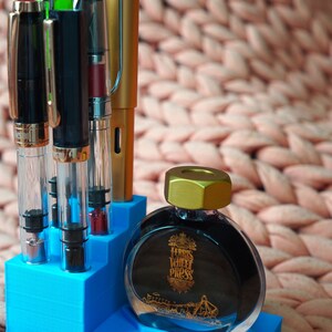 Puede incluir: Un portaplumas azul con cuatro plumas y una botella de tinta Ferris Wheel Press. La botella de tinta es de vidrio transparente con una tapa dorada y una etiqueta negra con letras doradas. La etiqueta dice "Ferris Wheel Press".